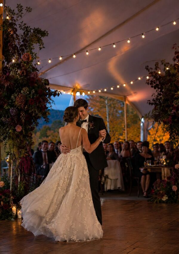 A bride and groom share their first dance under a canopy adorned with string lights and floral arrangements in rich fall wedding colors, surrounded by seated guests, with warm, ambient evening light in the background.
