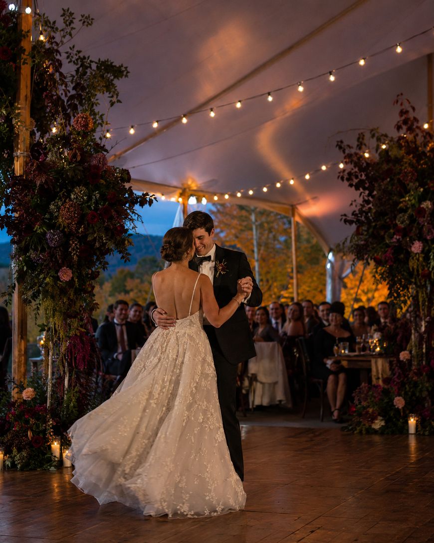 A bride and groom share a romantic first dance under string lights at an outdoor wedding reception, surrounded by lush floral arrangements and seated guests, with autumn trees in the background.