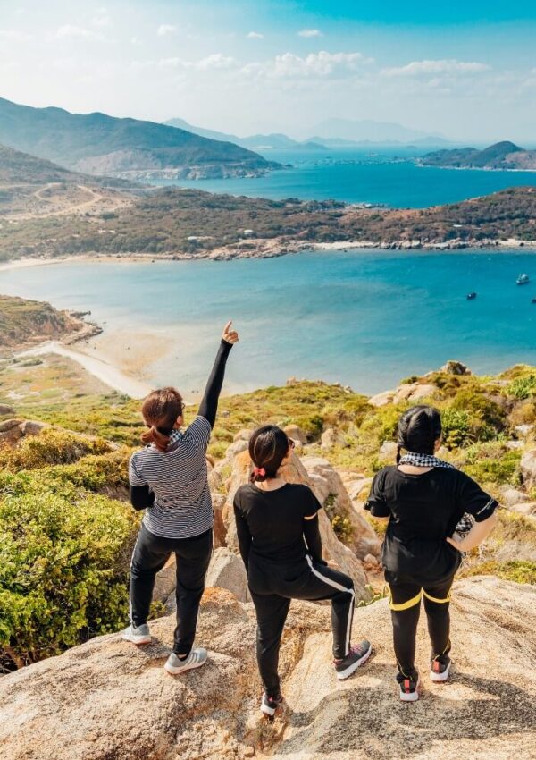 Three people in athletic wear stand on a rocky hilltop, overlooking a scenic blue bay surrounded by mountains. One person points towards the water, while the others admire the panoramic view below.