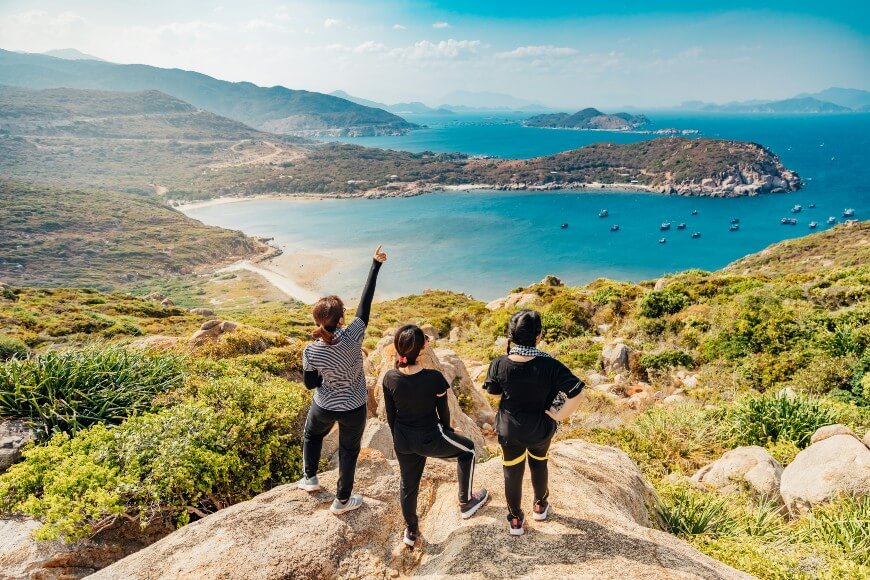 Three people in athletic clothing stand on rocks overlooking a scenic coastal bay with blue water, distant mountains, and boats. One person points toward the view. The sky is clear and sunny.