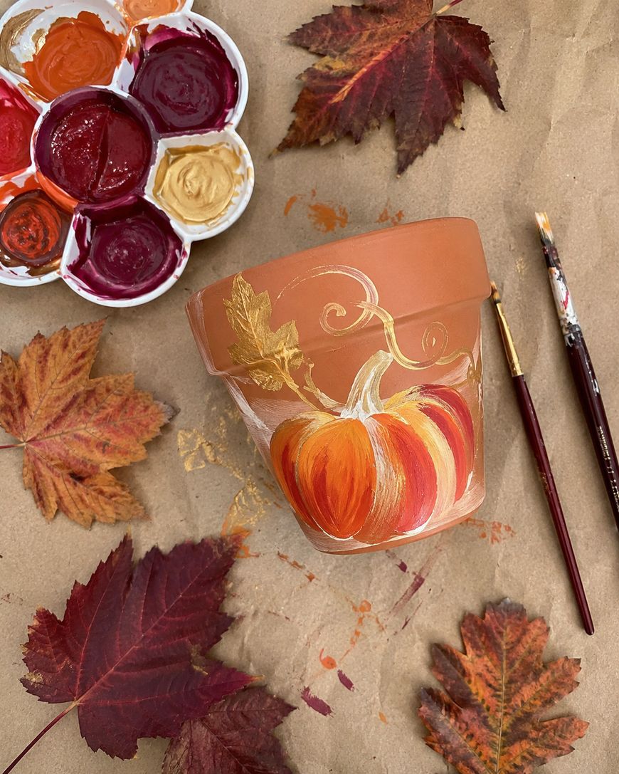 A terracotta pot painted with an orange pumpkin, golden leaves, and swirls sits on brown paper. Red and orange autumn leaves, paintbrushes, and a paint palette with red and gold paints surround the pot.