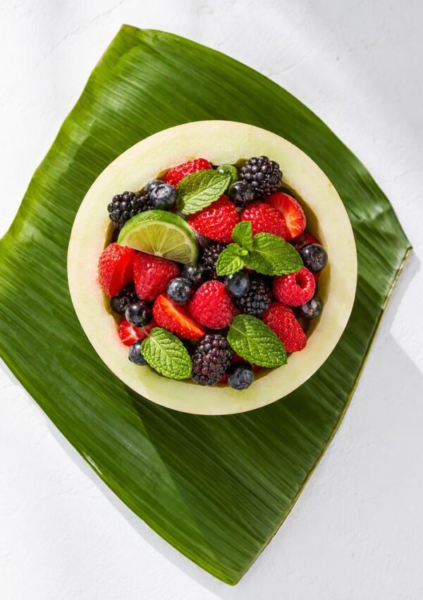A creative fruit cup idea: a melon bowl filled with strawberries, raspberries, blackberries, blueberries, lime slices, and mint leaves, placed on a large green leaf against a white background—perfect for party fruit recipes.