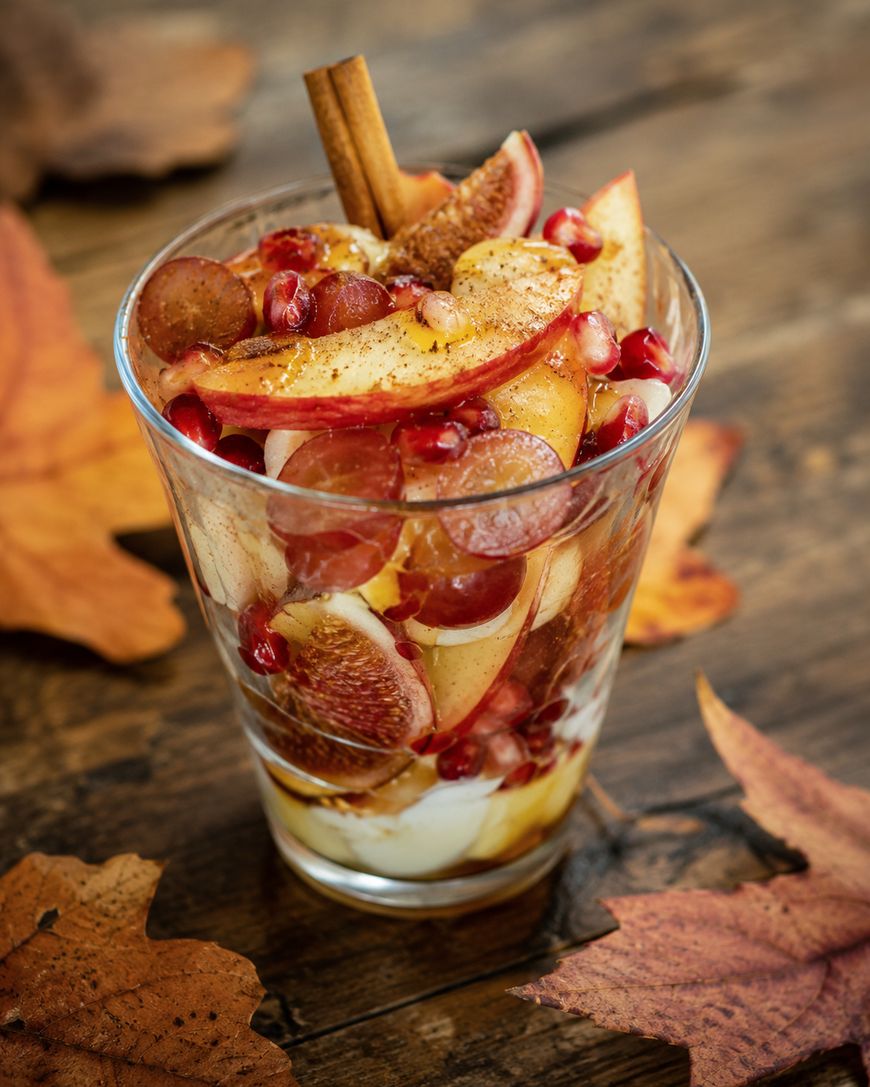 A glass filled with layers of sliced apples, grapes, figs, pomegranate seeds, yogurt, and honey, topped with a cinnamon stick, sits on a wooden table surrounded by autumn leaves.