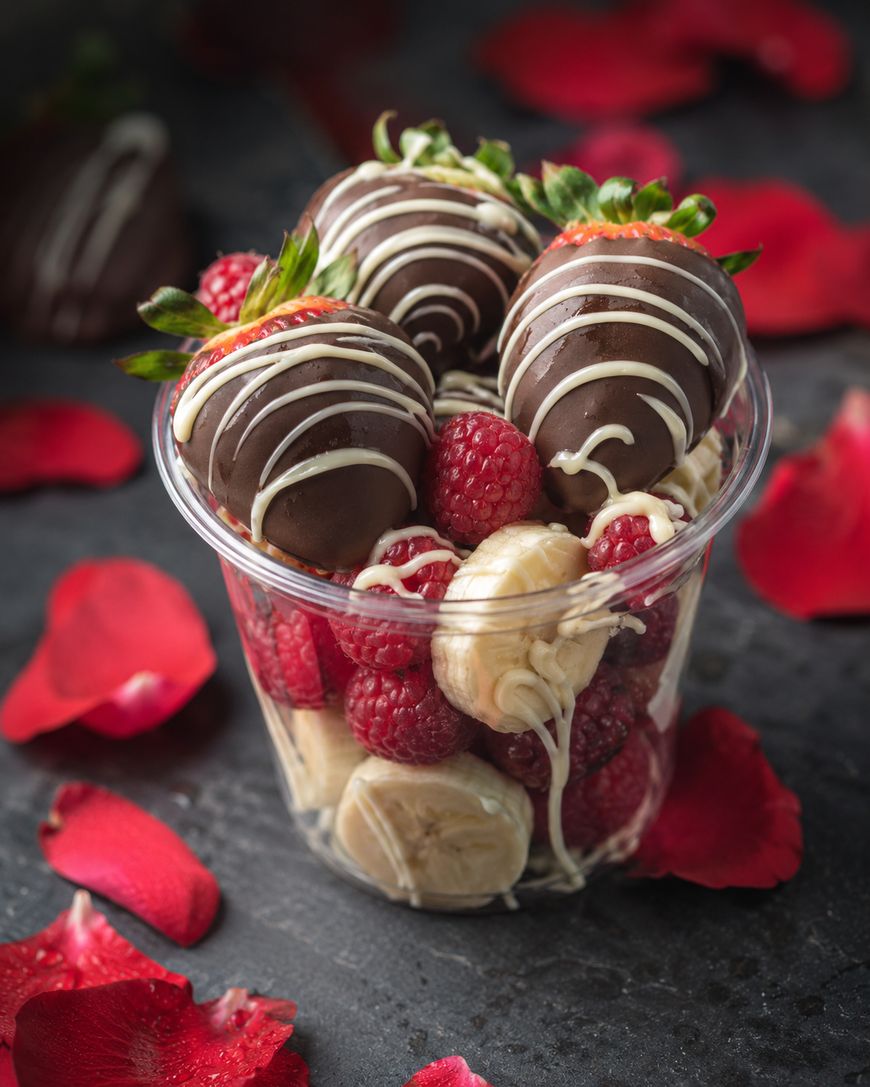 A clear cup filled with chocolate-covered strawberries, raspberries, and banana slices, surrounded by scattered red rose petals on a dark surface.