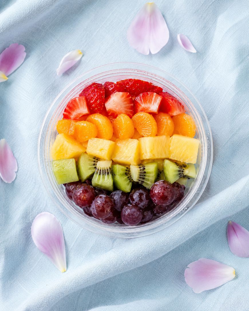 A clear bowl filled with neatly arranged rows of strawberries, orange slices, pineapple, kiwi, and grapes, creating a rainbow effect. The bowl sits on a light blue cloth with scattered pink flower petals.