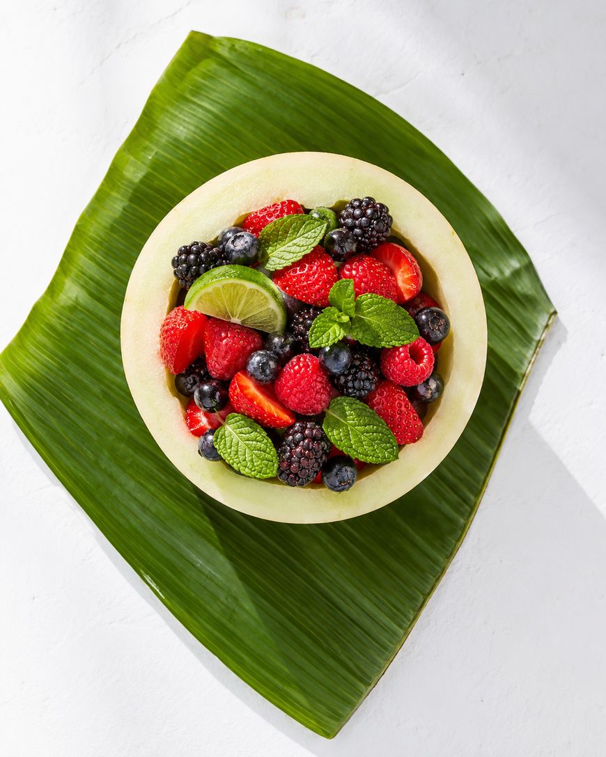 A bowl made from a hollowed melon filled with strawberries, blackberries, raspberries, blueberries, lime slices, and mint leaves, placed on a large green leaf on a white background.
