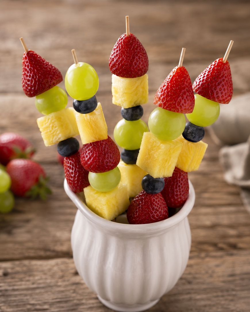 Skewers with strawberries, green grapes, pineapple chunks, blueberries, and red grapes are arranged in a white cup on a wooden table.