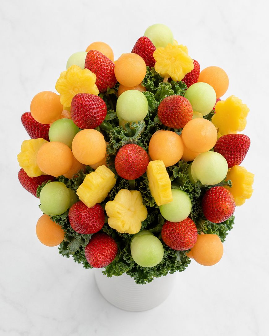 A bouquet arrangement of strawberries, cantaloupe balls, honeydew melon balls, and pineapple flowers, set on a bed of leafy greens in a white container, viewed from above.