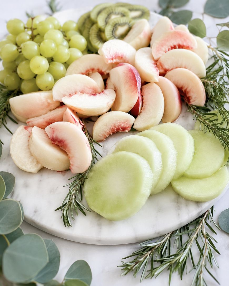 A marble platter topped with sliced white peaches, green honeydew melon, kiwi, and a bunch of green grapes, garnished with sprigs of rosemary and eucalyptus leaves.