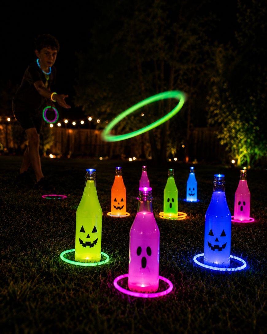 A child plays a nighttime ring toss game using glowing rings and neon bottles decorated with jack-o’-lantern faces, set up on grass with string lights in the background.