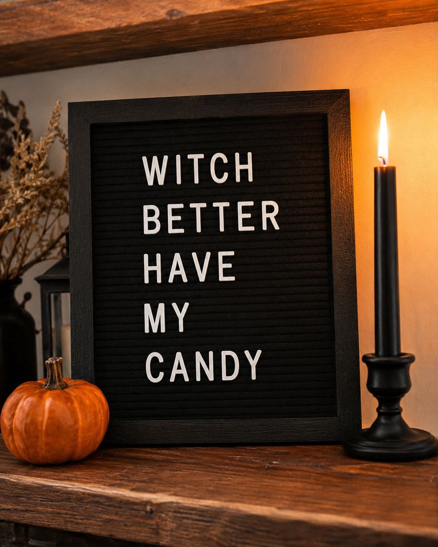 A black letter board with white letters reads WITCH BETTER HAVE MY CANDY. It sits on a wooden shelf next to a small pumpkin, a lit black candle, and dried flowers, creating a Halloween-themed scene.