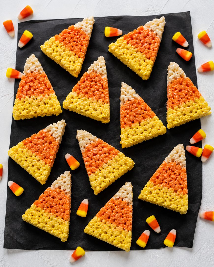 Rice Krispie treats decorated to look like candy corn with yellow, orange, and white layers are arranged on a black square napkin, surrounded by actual candy corn pieces on a white surface.