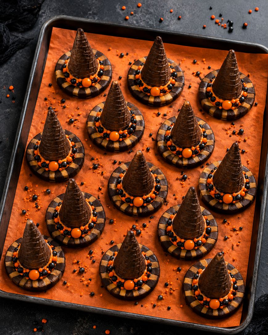 A baking tray holds 12 cookies decorated to look like witch hats, with chocolate cone tops, round checkered cookie bases, orange candy, and black and orange sprinkles, all on orange parchment paper.