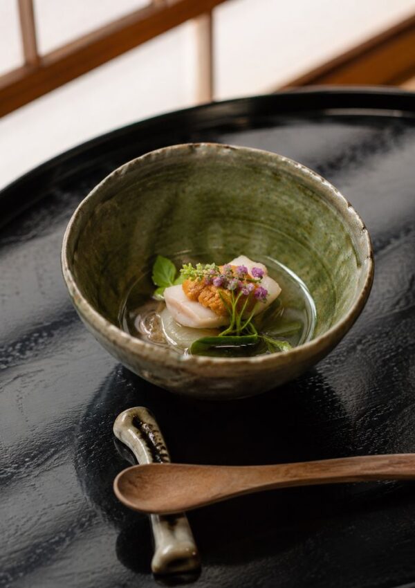 A ceramic bowl with a delicate dish, possibly scallop with edible flowers and herbs, sits on a black tray beside a wooden spoon—perfect for savoring quiet moments after the wedding or dreaming of the best honeymoon destinations.