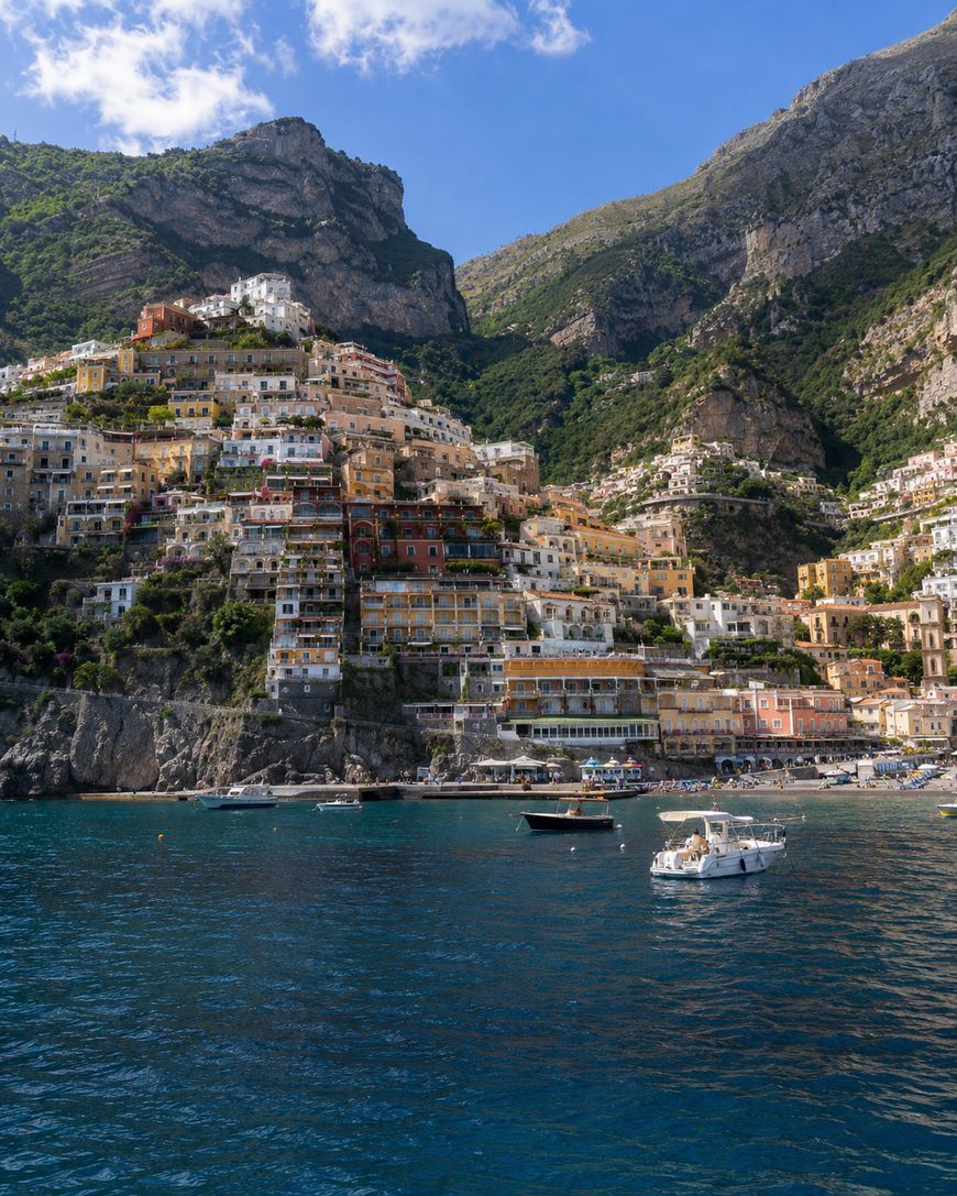 Colorful buildings cling to steep cliffs along the coast of Positano, Italy, with boats floating on the deep blue sea and lush green mountains rising in the background under a partly cloudy sky.