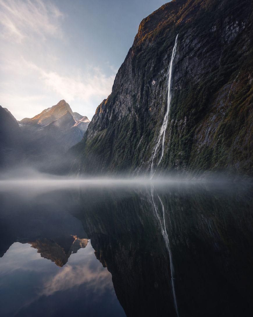 A tall waterfall cascades down a steep, green mountainside, reflected in the still water below. Mist hovers above the lake, with rugged peaks in the background under a partly cloudy sky.