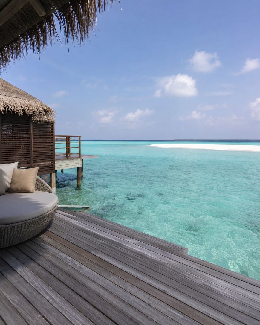 A cozy outdoor lounge chair sits on a wooden deck over clear turquoise water, next to a thatched-roof villa, with a bright sky and distant white sandbank in the background.