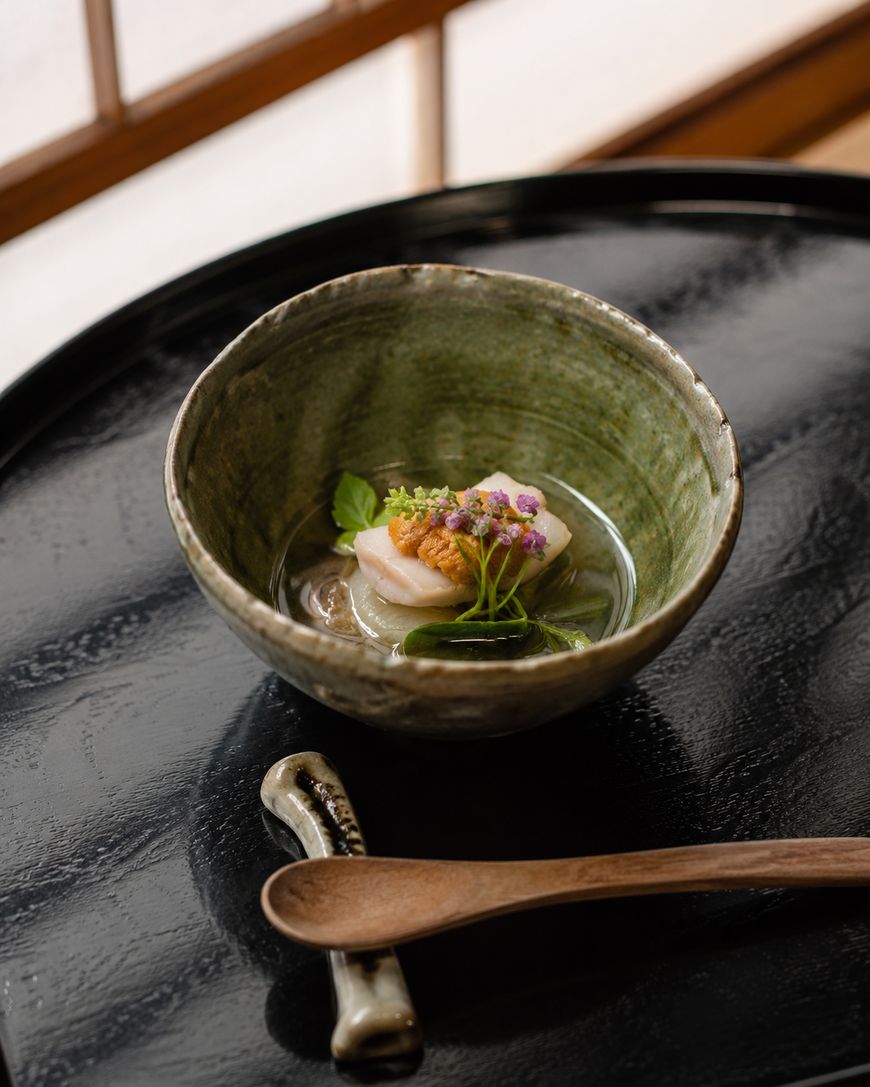 A ceramic bowl with elegantly plated scallops, garnished with microgreens and edible flowers, sits on a black tray next to a wooden spoon. Soft natural light streams in from a window in the background.