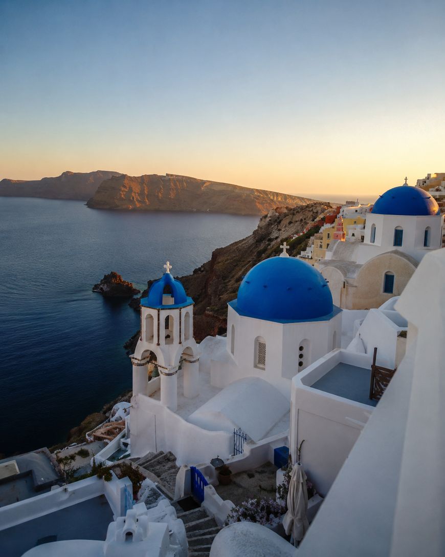 White buildings with blue domes overlook the sea at sunset in Santorini, Greece. The calm water and distant island are visible under a clear sky, with warm golden light illuminating the scene.