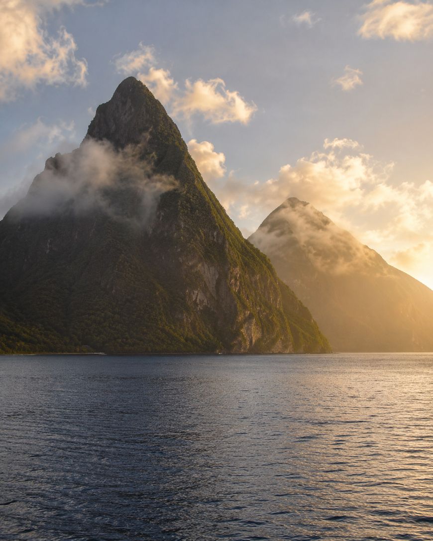 Two lush, steep mountain peaks rise above the water, partially shrouded in mist and bathed in golden sunlight, with a calm body of water in the foreground.