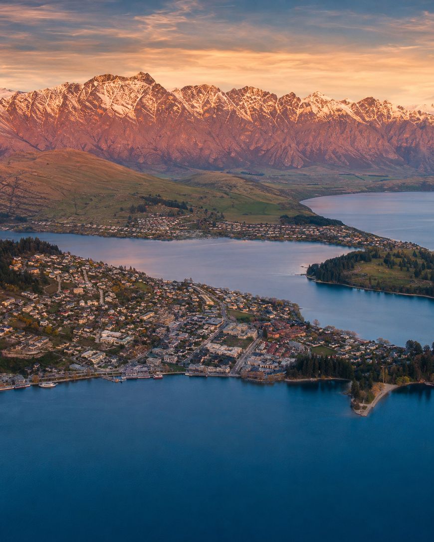 Aerial view of a lakeside town with houses clustered along the shore, surrounded by deep blue water and backed by snow-capped mountains under a golden sunset sky.