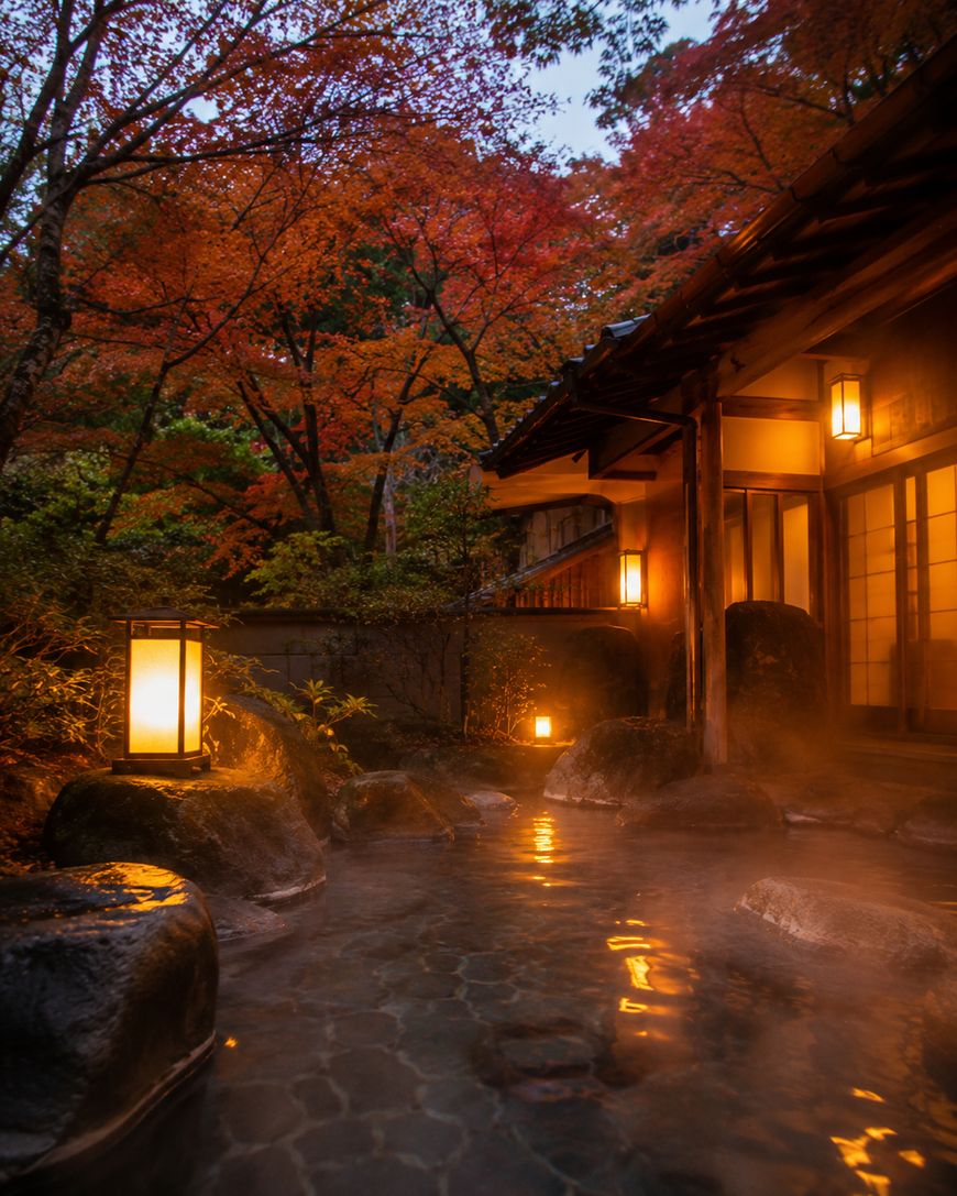 Outdoor hot spring bath surrounded by rocks and trees with autumn foliage, soft lanterns glow warmly beside a traditional Japanese building at dusk, creating a tranquil, inviting atmosphere.