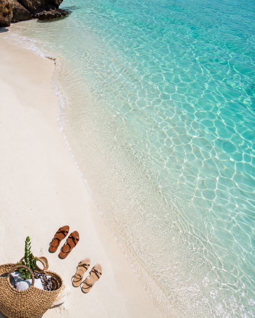 A woven beach bag and three pairs of sandals rest on soft white sand beside clear turquoise water under bright sunlight at a peaceful beach.