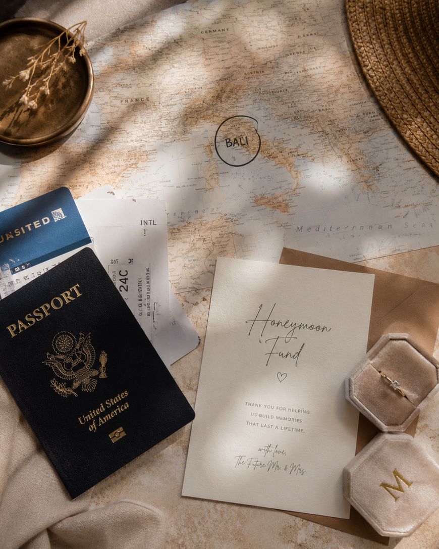 Flat lay of a travel-themed setup with a map, U.S. passport, airline boarding pass, straw hat, wedding rings in boxes, and a “Honeymoon Fund” card, suggesting a honeymoon trip to Bali.