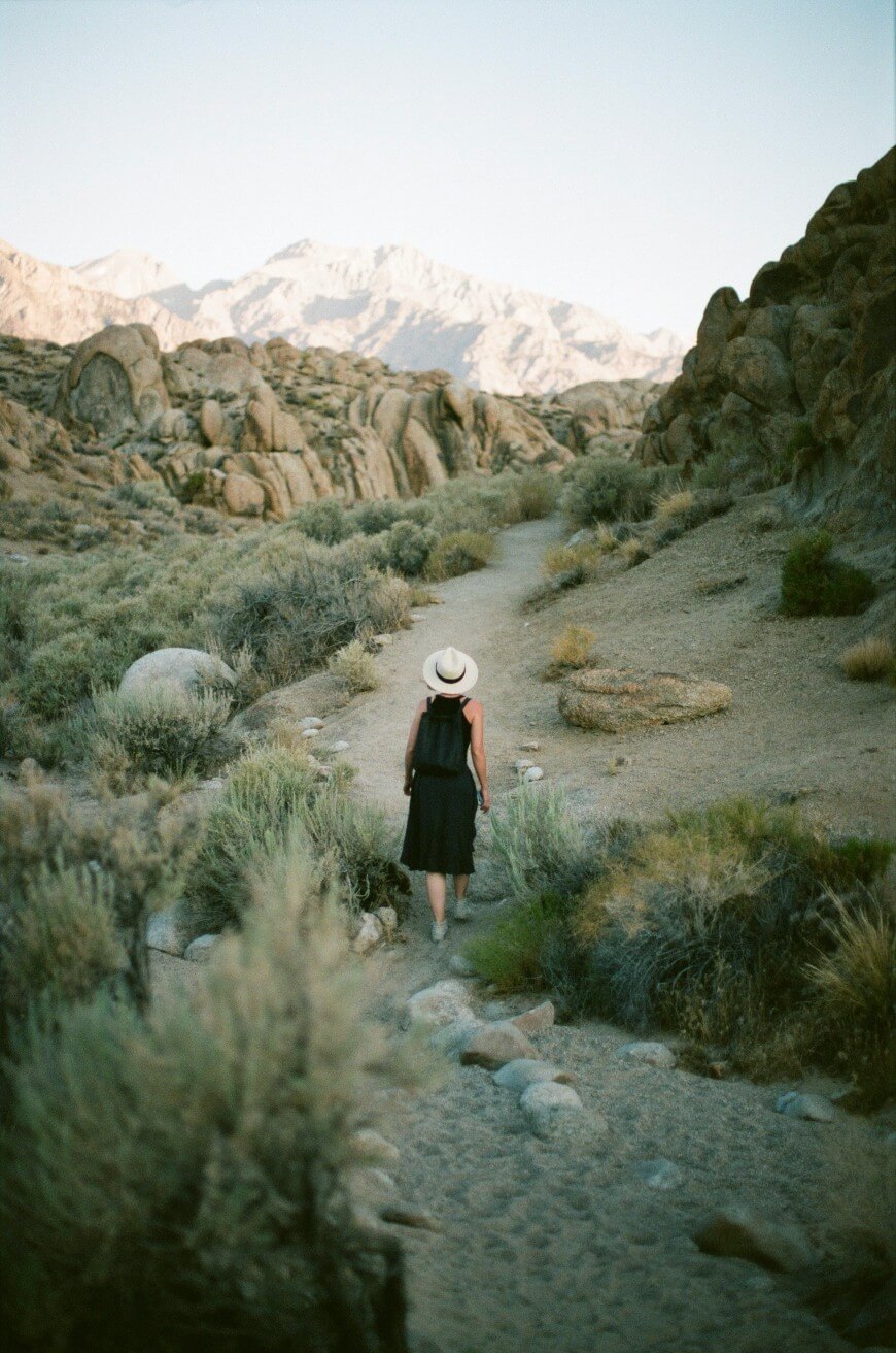 A person wearing a black dress and a white hat walks alone on a narrow dirt trail through a rocky, desert landscape with mountains in the background.