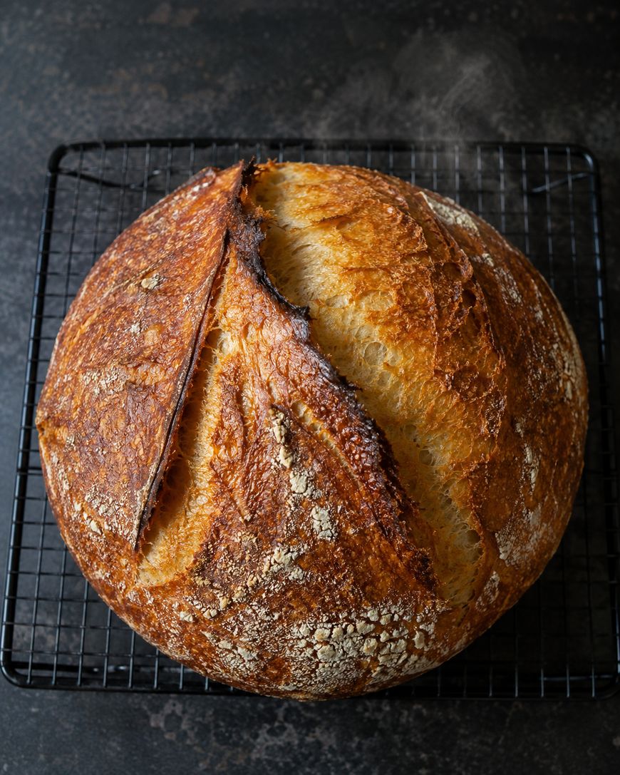 A round loaf of rustic sourdough bread with a golden, crusty top rests on a black wire cooling rack against a dark background. The bread has a deep slash on top and a textured, flour-dusted surface.