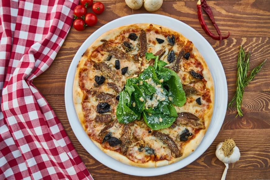 A pizza topped with mushrooms, black olives, and fresh spinach sits on a white plate. The table is set with a red checkered cloth, cherry tomatoes, garlic, rosemary, and mushrooms.