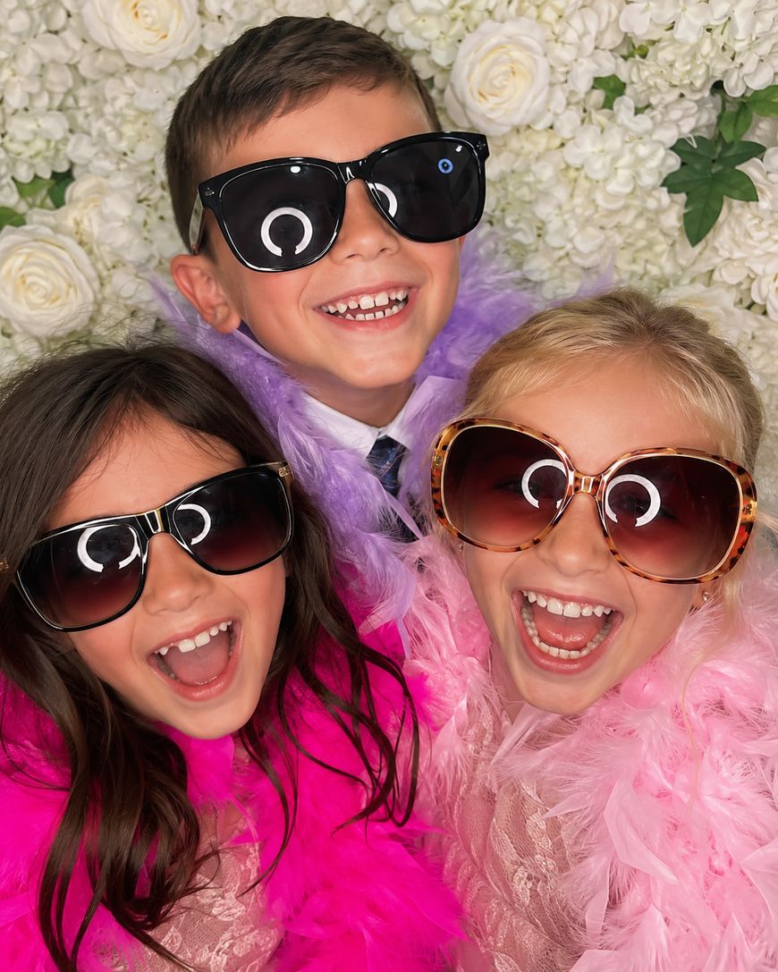 Three smiling children wearing large sunglasses and colorful feather boas pose together in front of a white flower backdrop, looking joyful and playful.