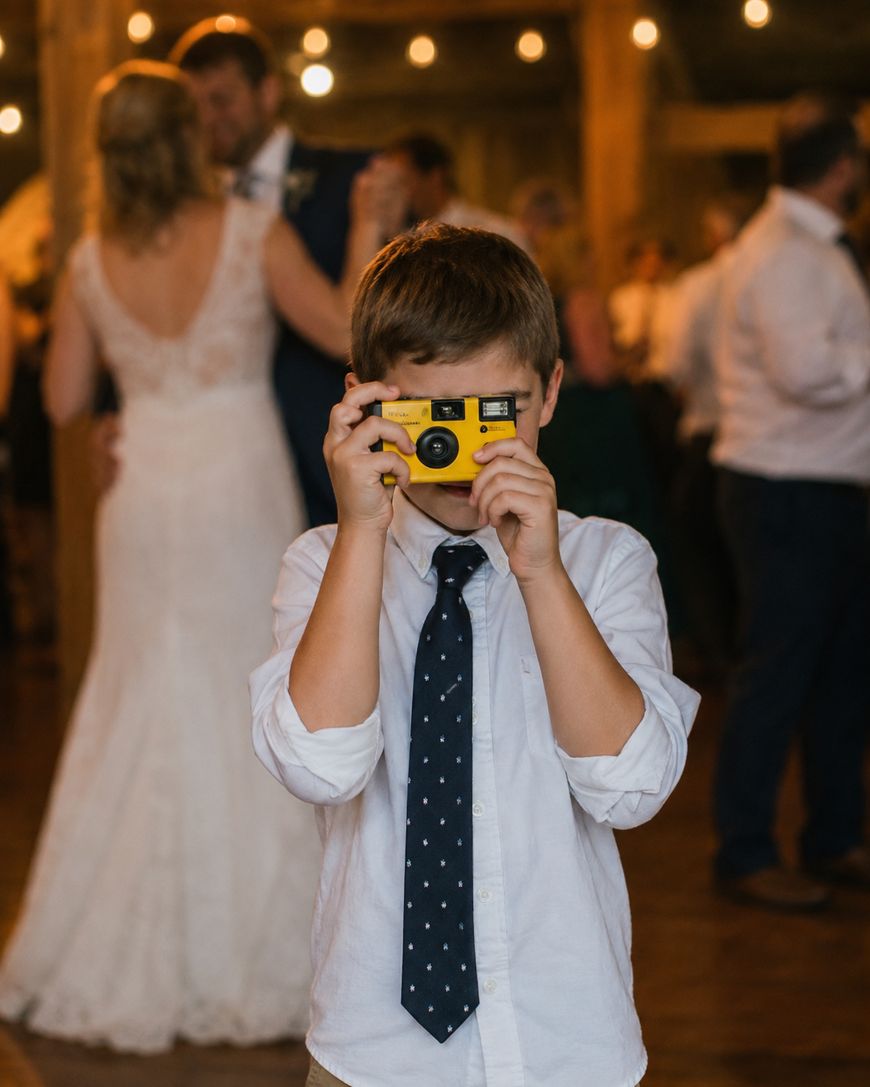 A young boy in a white shirt and polka-dot tie holds up a yellow toy camera, pretending to take a photo at a wedding reception. Adults, including a bride in a white dress, dance in the softly-lit background.