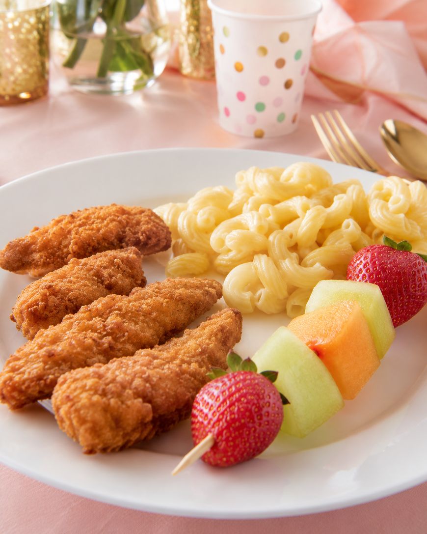 A white plate with three breaded chicken tenders, a serving of macaroni and cheese, and a fruit skewer with strawberry, cantaloupe, and honeydew melon. A paper cup and gold utensils are in the background.