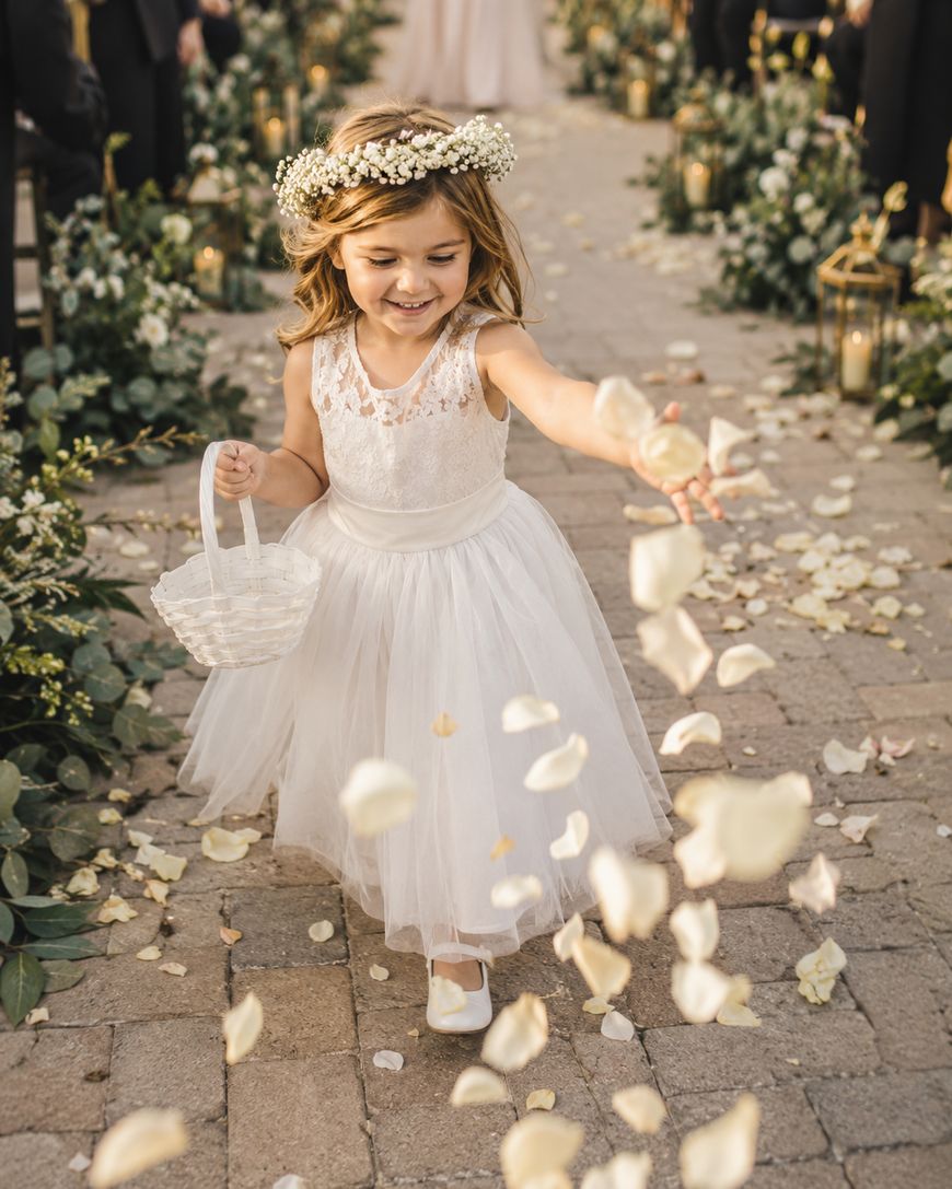 A young flower girl in a white dress and floral crown smiles as she scatters rose petals from a basket along an outdoor wedding aisle lined with flowers and lanterns.