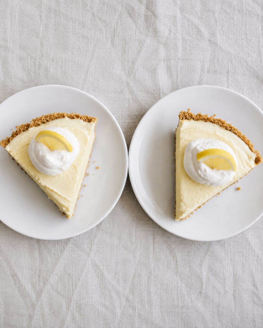 Two plates, each with a slice of creamy pie topped with a dollop of whipped cream and a small lemon wedge, are placed side by side on a light-colored, textured tablecloth.