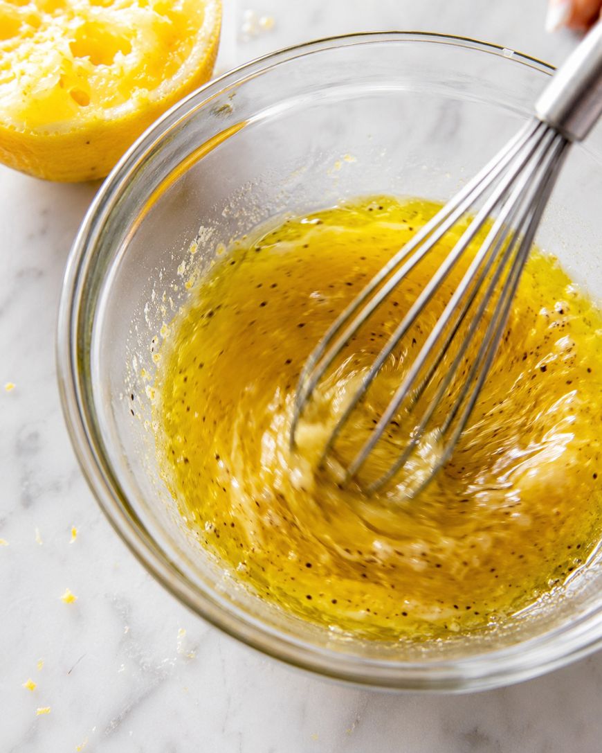 A metal whisk mixing a yellow, seasoned liquid in a glass bowl, with a halved lemon nearby on a marble surface.