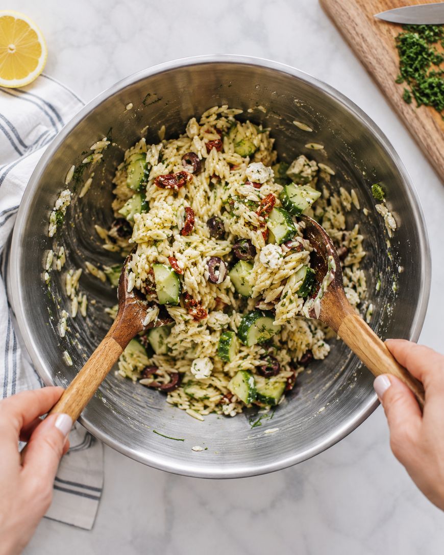 A person mixing orzo pasta salad with cucumbers, feta, olives, and herbs in a large metal bowl using wooden salad servers; a cutting board and lemon are nearby on a marble surface.