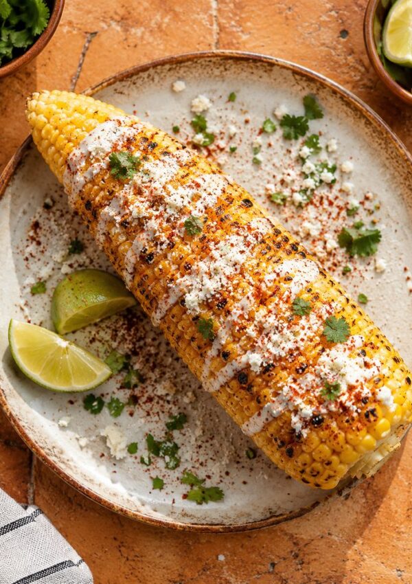 A grilled ear of corn on a plate, topped with creamy white sauce, crumbled cheese, chili powder, and cilantro, served with lime wedges and garnished with extra cilantro for a tangy finish. Inspired by our Lime Crema Recipe.