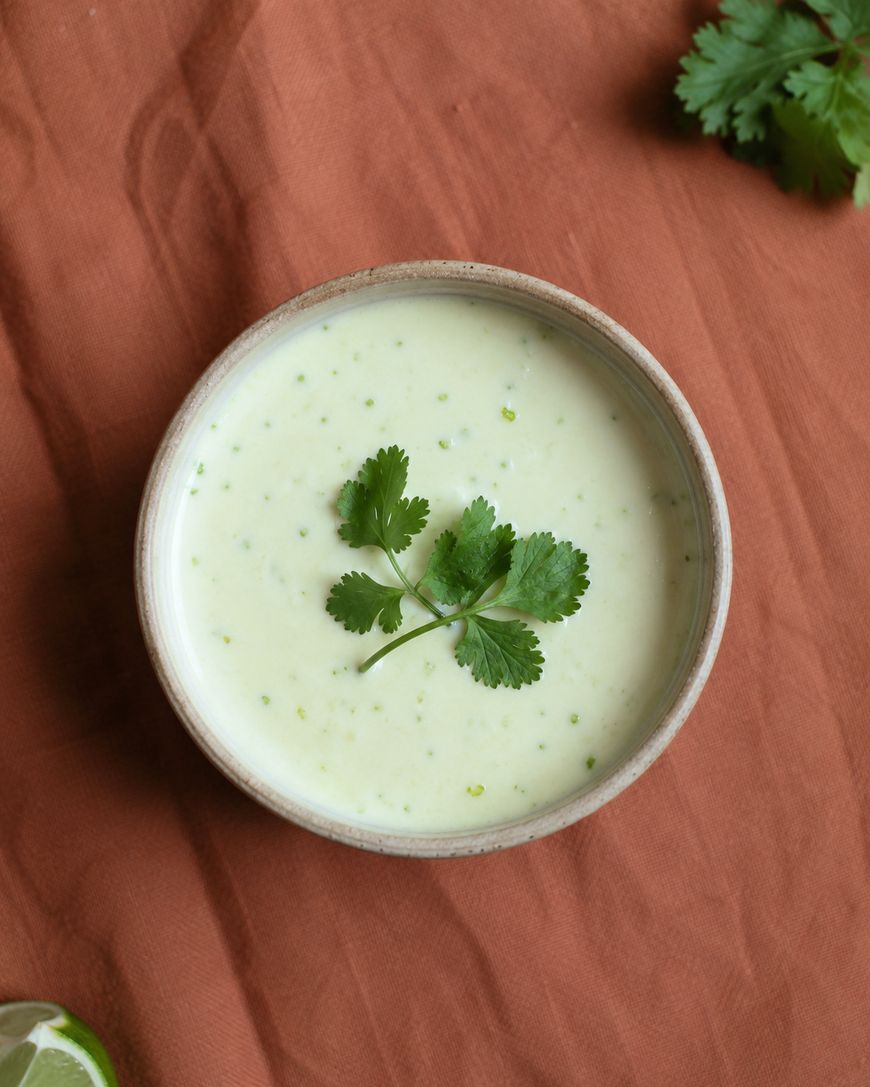 A bowl of creamy green sauce garnished with fresh cilantro sits on a rust-colored cloth, with extra cilantro and a lime wedge nearby.