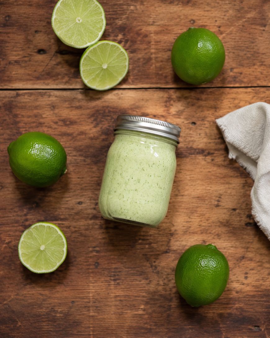 A jar of creamy green sauce sits on a wooden surface surrounded by whole and halved limes, with a white cloth in the corner.