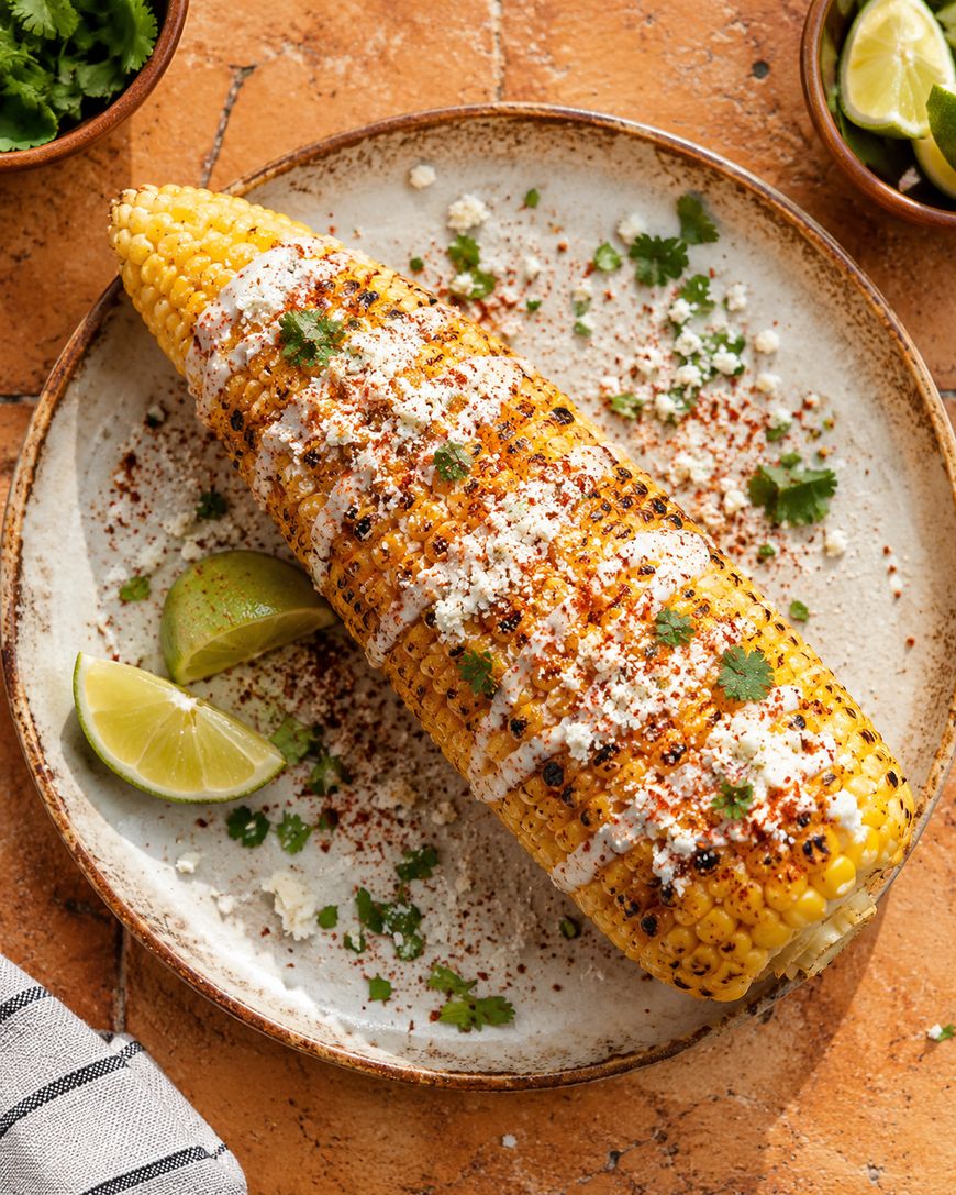A grilled ear of corn on a plate, topped with creamy sauce, crumbled cheese, chili powder, and cilantro, with lime wedges on the side. Small bowls of herbs and lime are in the background.