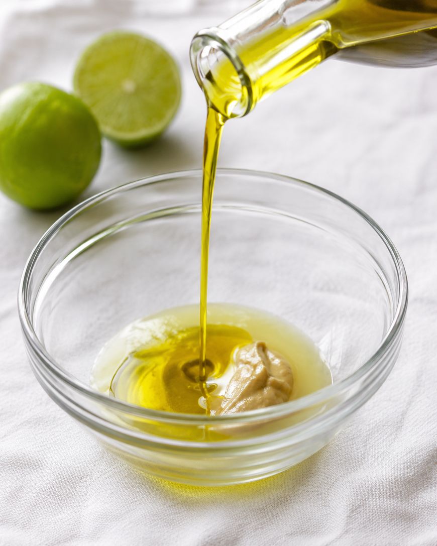 Olive oil is being poured into a glass bowl containing mustard, with two limes—one whole and one halved—in the background on a white cloth surface.