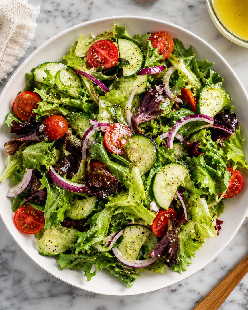 A fresh salad with mixed greens, sliced cucumbers, cherry tomatoes, and thinly sliced red onions, lightly dressed and served in a white bowl on a marble surface.