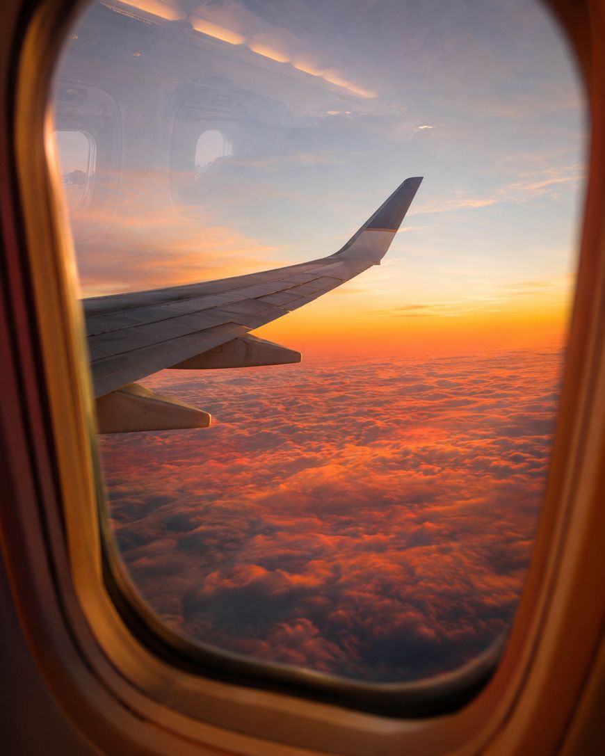 A view from an airplane window showing the wing and a vibrant orange sunset over a blanket of clouds. The sky is filled with warm colors, creating a peaceful and scenic atmosphere.