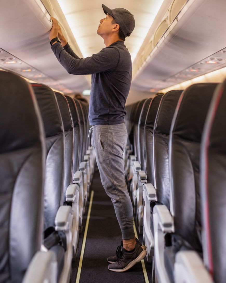 A person in casual clothes and a cap stands in an airplane aisle, reaching up to place or retrieve an item from the overhead compartment. The airplane seats are empty.