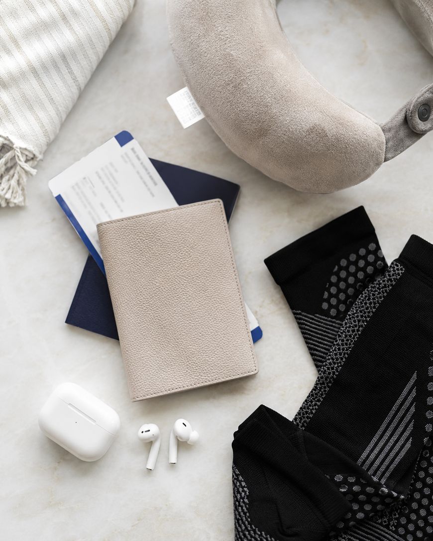 Flat lay of travel essentials: a neck pillow, striped fabric, airline tickets, passport with a beige cover, black patterned socks, and wireless earbuds with their case on a light surface.