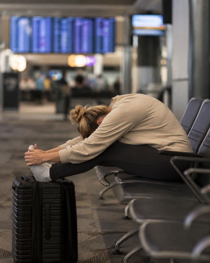 A woman sits in an airport terminal, stretching with her legs resting on a suitcase and her head down on her knees. She appears tired, with a departure board visible in the background.