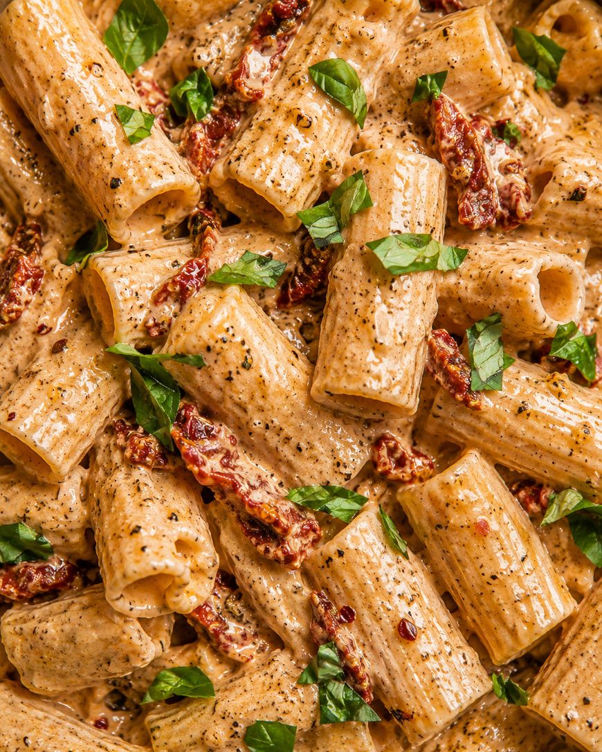 Close-up of creamy rigatoni pasta mixed with sun-dried tomatoes, black pepper, and fresh basil leaves. The sauce appears rich and the pasta is generously coated, with herbs sprinkled on top for garnish.