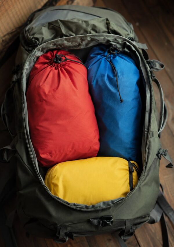 An open backpack containing three stuff sacks—one red, one blue, and one yellow—neatly packed inside. This essential piece of outdoor gear rests on a wooden floor, ready for your next outdoor travel adventure.
